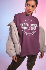 Model wearing a purple 'Hyperfixation World Tour' graphic t-shirt, styled with a beige jacket and casual pants.