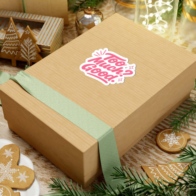Gift box adorned with a colorful 'Too Much Good!' kiss-cut sticker, surrounded by holiday decor.