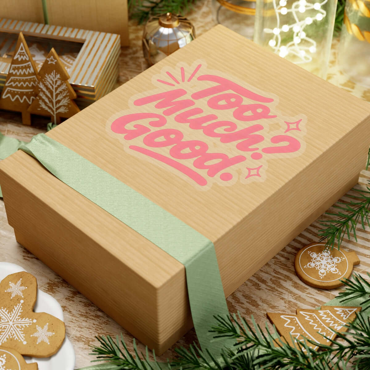 Gift box with the phrase 'Too Much? Good!' in pink, decorated with green ribbon and holiday ornaments.