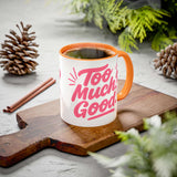 Colorful coffee mug with the phrase 'Too Much Good' on a wooden cutting board, surrounded by pinecones.