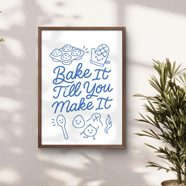 Framed motivational poster with 'Bake It Till You Make It' text in a room with plants and wooden floor.