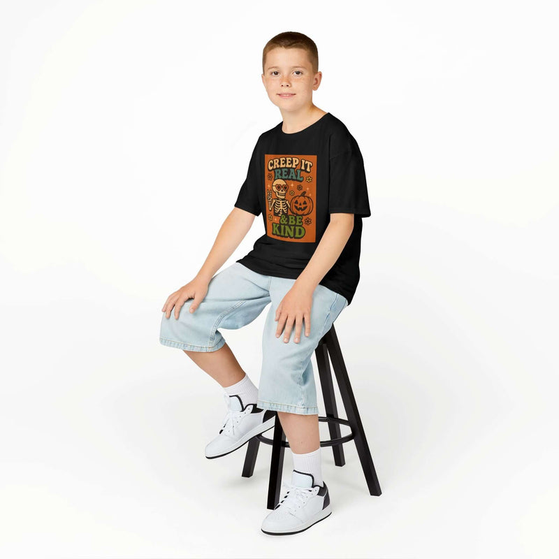 Boy sitting on stool wearing Global Halloween Kids Tee featuring 'Creep It Real & Be Kind' design.