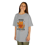 Smiling child wearing a gray tee with cartoon cat and 'YOU MATTER' text, promoting positivity and self-love.