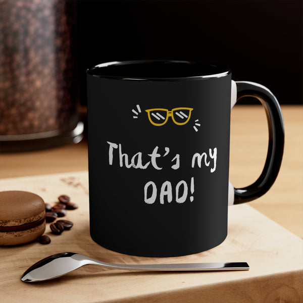 Black mug with 'That's my DAD!' text on a wooden surface with coffee beans and a spoon.