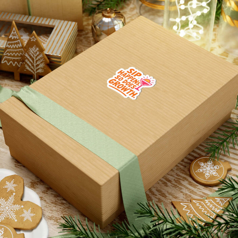 Gift box featuring 'Sip Happens' sticker, surrounded by holiday decorations and greenery for joyful vibes.