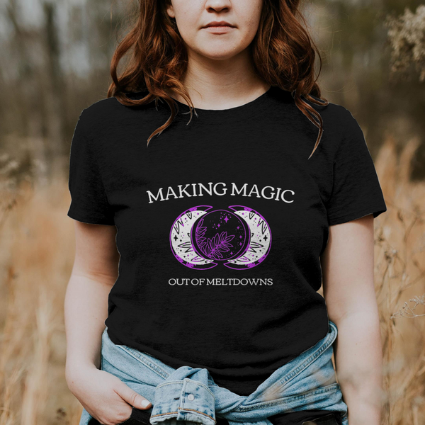 Person wearing a black t-shirt with 'Making Magic Out of Meltdowns' design in a field.